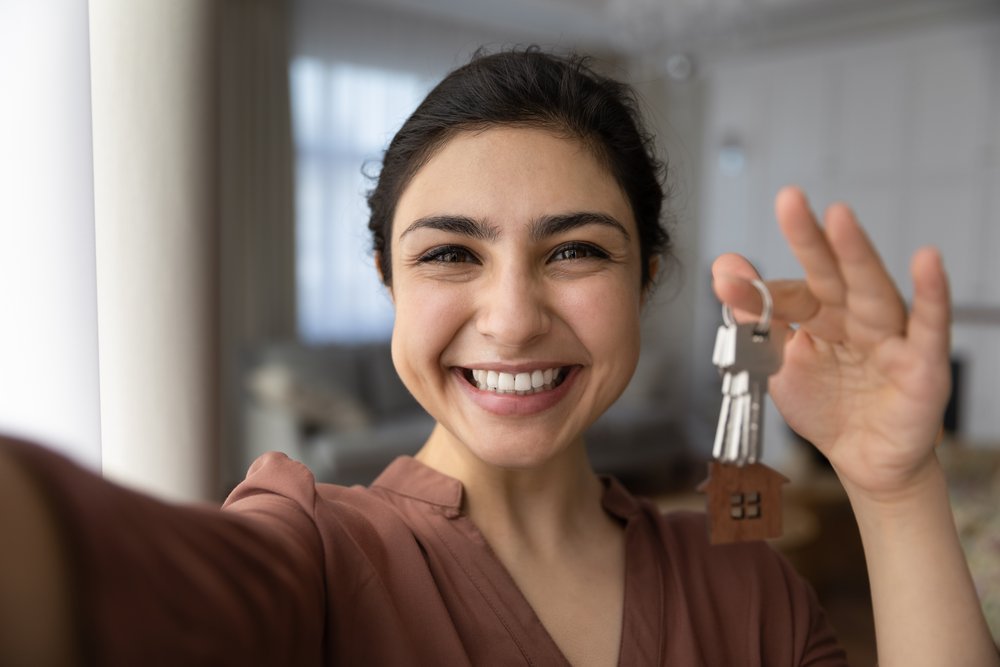 Close up self-portrait picture of happy Indian woman feel excited show keys to new home. Selfie of smiling mixed race ethnicity female renter or tenant celebrate moving or relocation to own house.