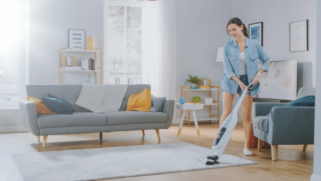 Young Woman in Jeans Shirt and Shorts is Vacuum Cleaning a Carpet in a Bright Cozy Room at Home. She Uses a Modern Cordless Vacuum. She's Happy and Cheerful.