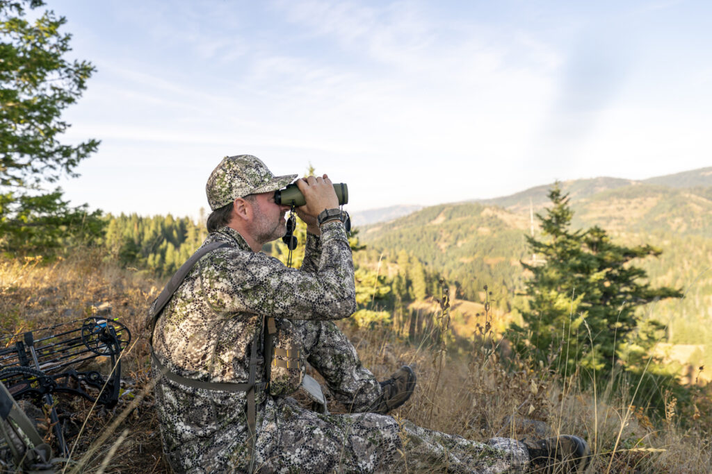 A bowhunter sits on a mountain peak and looks through binoculars while tracking wild game in the forested wilderness. A crossbow is lying on the ground behind the man.