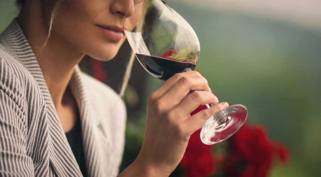 Closeup of unrecognizable adult woman holding a glass of red wine and smelling it before tasting. She's standing outdoors on summer afternoon. Blurry gras and red flowers in background. Toned image.