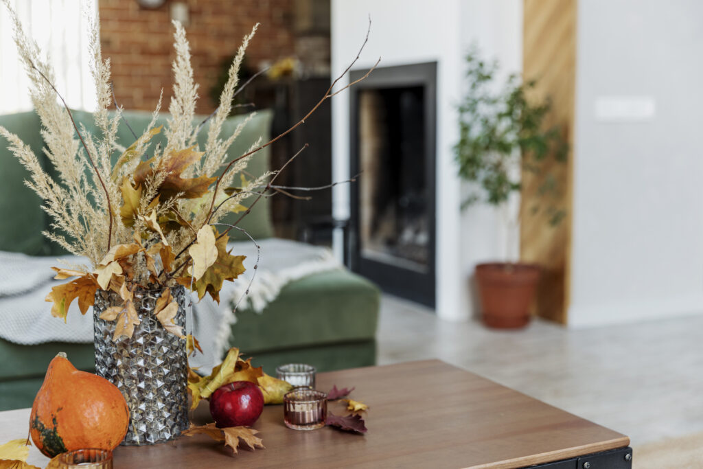 Autumn time decorated home interior. Yellow flowers bouquet in glass vase on wooden stylish table, pumpkin, red apples, leaves, candles, blanket. Green sofa, brick wall and fireplace on background