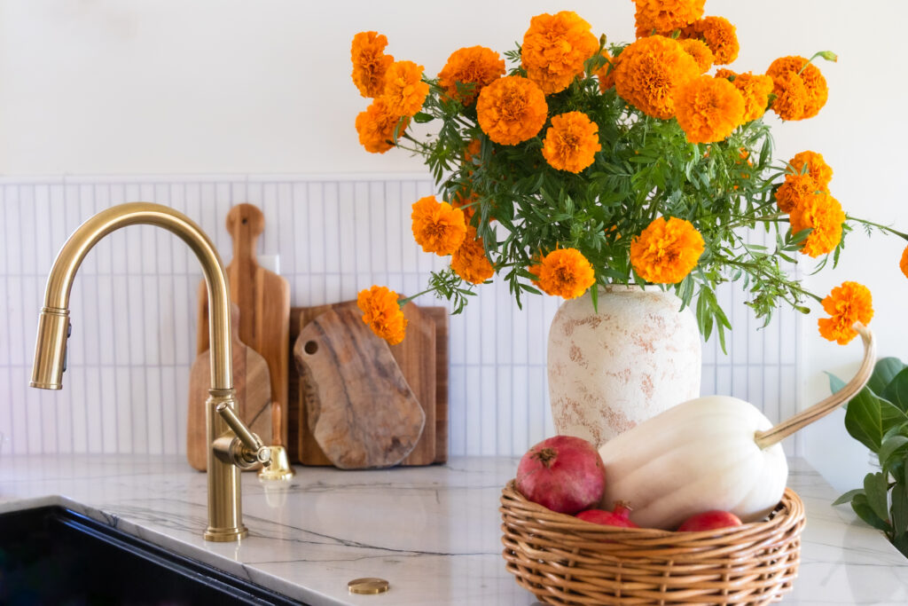 Fall kitchen setting with marigolds. A woven basket with pomegranate and squash. Large white vase full of flowers. Marble backsplash and countertop.